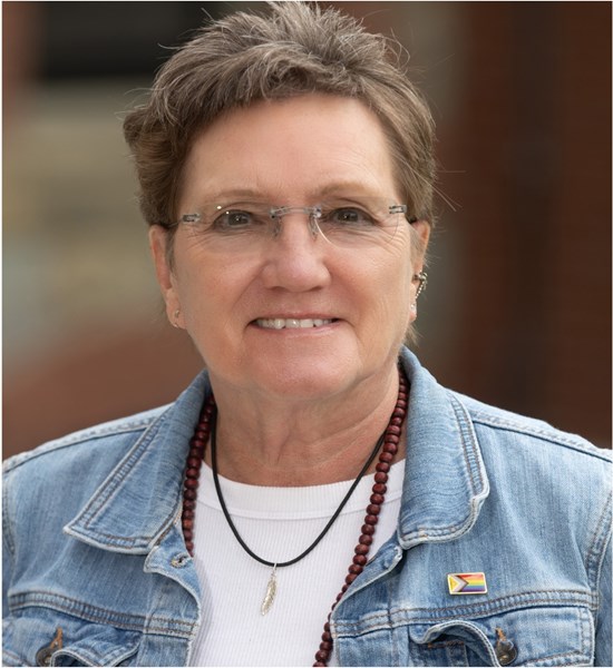 Professional headshot of Michele Forinash wearing glasses, a light blue denim jacket over a white shirt, and two necklaces. The portrait is set against a softly blurred indoor background with neutral tones.